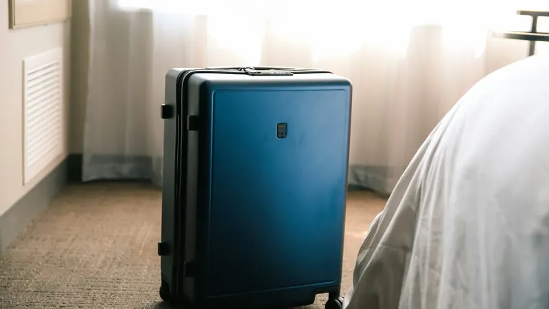 Blue hard-shell suitcase beside a hotel bed in morning light